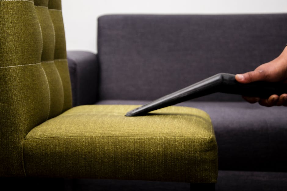 A close-up image showing a hand operating a portable steam cleaning device on a beige car seat with a textured fabric surface. The device's nozzle is pressed onto the seat cushion, emitting steam for