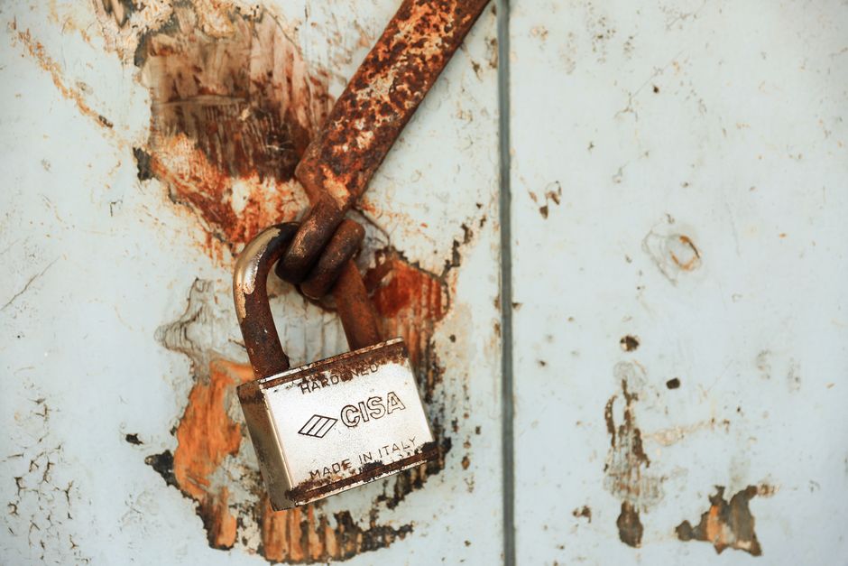 Close-up image of rusted padlocks with printed labels, attached to a metal latch, possibly part of a security or locking mechanism on a door or gate. The scene showcases weathered metal surfaces with